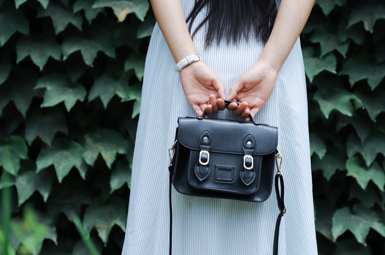 Home A woman in a striped dress holds a black leather handbag with lush green leaves in the background.